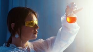 Woman in lab coat examining a vibrant liquid in a flask under lab lights.